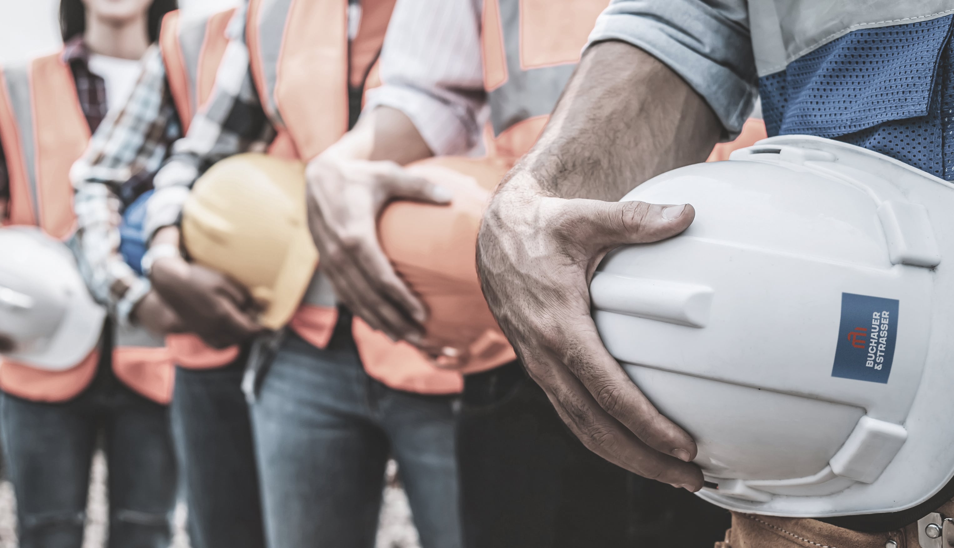 Eine Gruppe von Bauarbeitern trägt Sicherheitswesten und hält Bauhelme in den Händen. Die Helme sind in verschiedenen Farben, darunter weiß und orange. Die Szene vermittelt Gemeinschaft und Teamarbeit im Bauwesen.